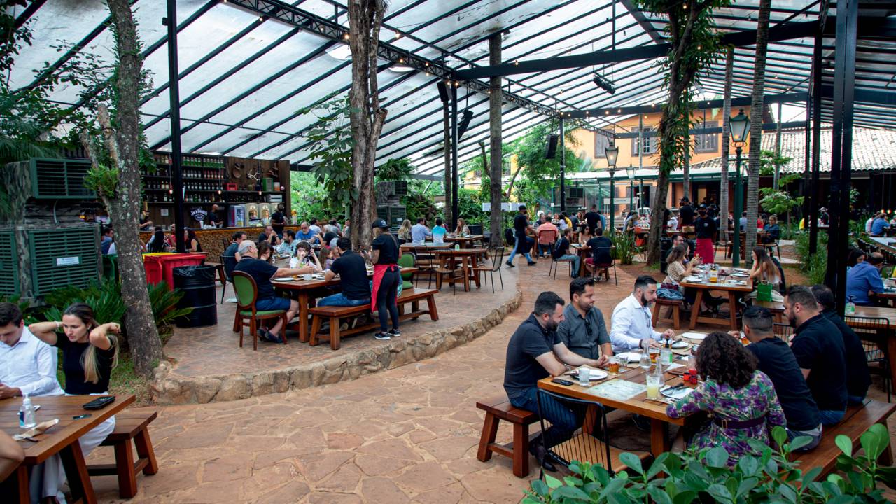 Salão de restaurante ao ar livre com teto transparente e árvores espaçadas entre as mesas.