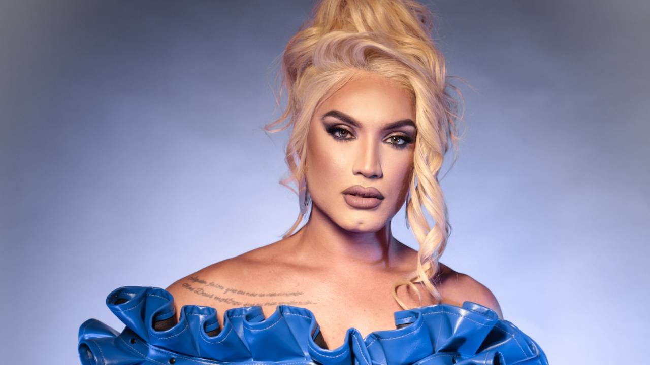 Drag queen loira veste traje azul com aparência de látex e babados na área do peito, além de luvas longas pretas.