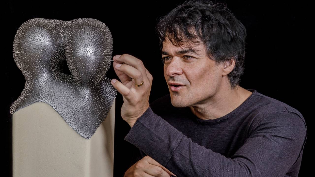 Homem branco grisalho posa concentrado colocando alfinete em escultura prateada