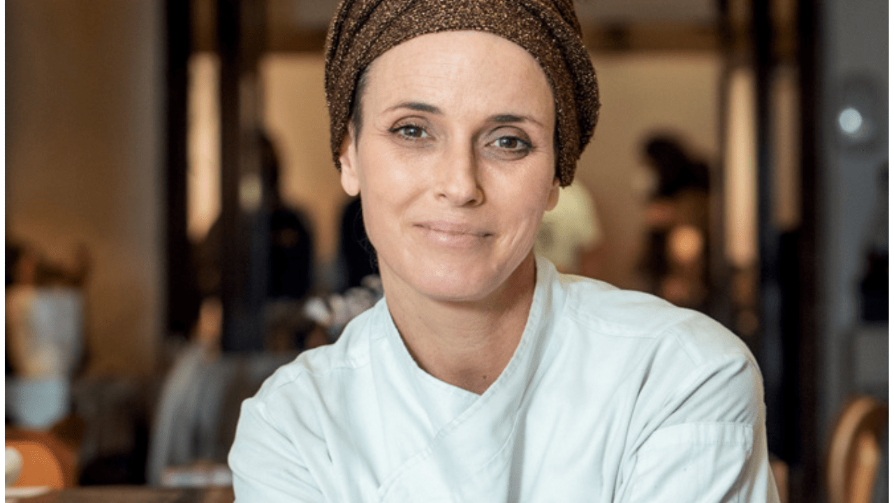 Close-up de mulher branca, chef de cozinha, com meio sorriso, de dólmã branco e lenço no cabelo.
