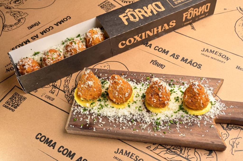 Forno Quatro coxinhas alinhadas sobre maionese em cima de prato de madeira cobertas por queijo ralado ao lado de caixa comprida retangular com coxinhas dentro.