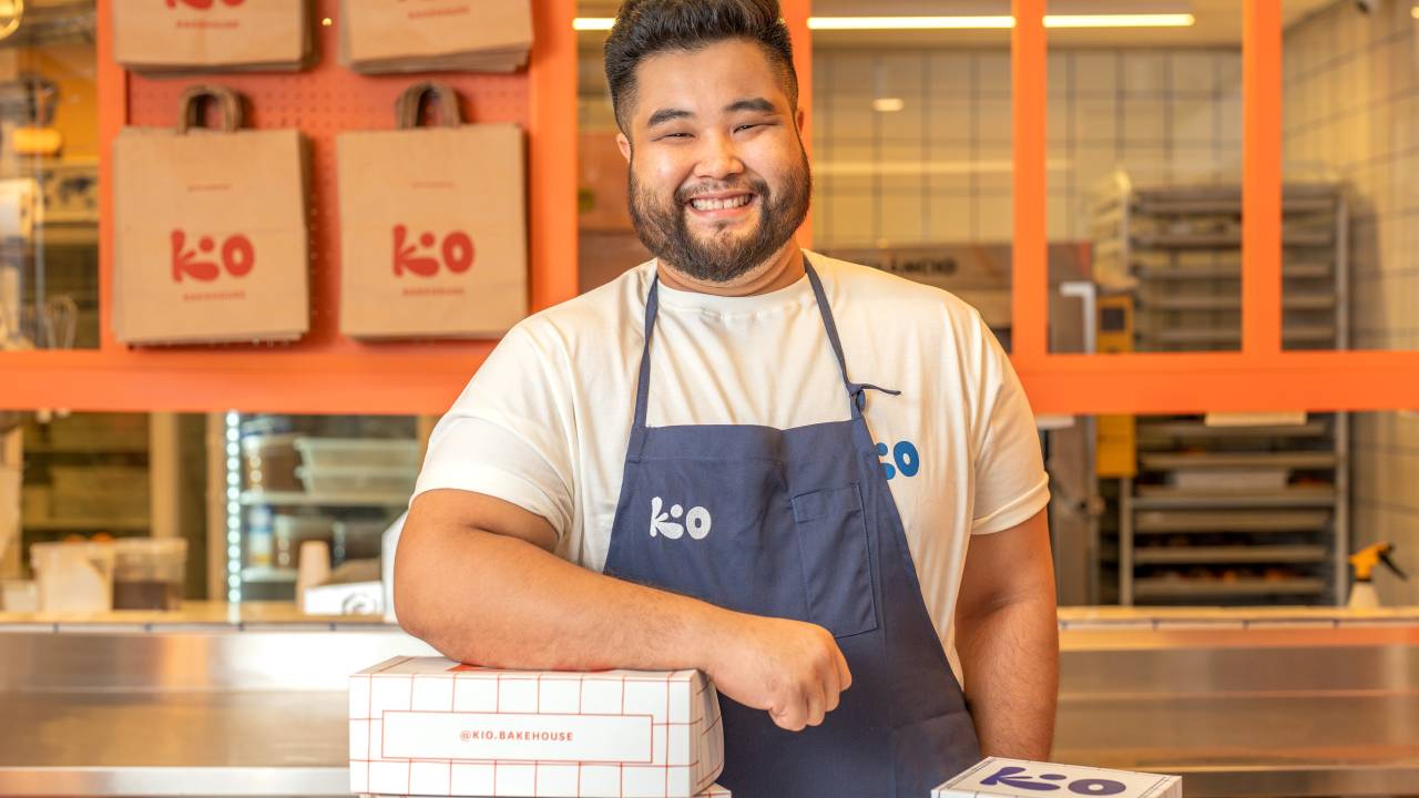 Chef de cozinha oriental de camiseta branca e avental azul por cima apoiando braço em caixa de entrega branca com cozinha industrial atrás.