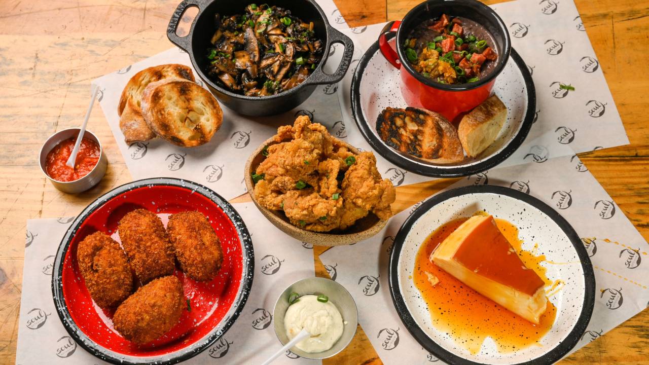 Mesa de madeira com diversos petiscos. À esquerda prato com bolinhos fritos, frango frito ao centro, pudim com calda no canto direito, caldinho de feijão em caneca no canto superior direito ao lado de panelinha de ferro com legumes.