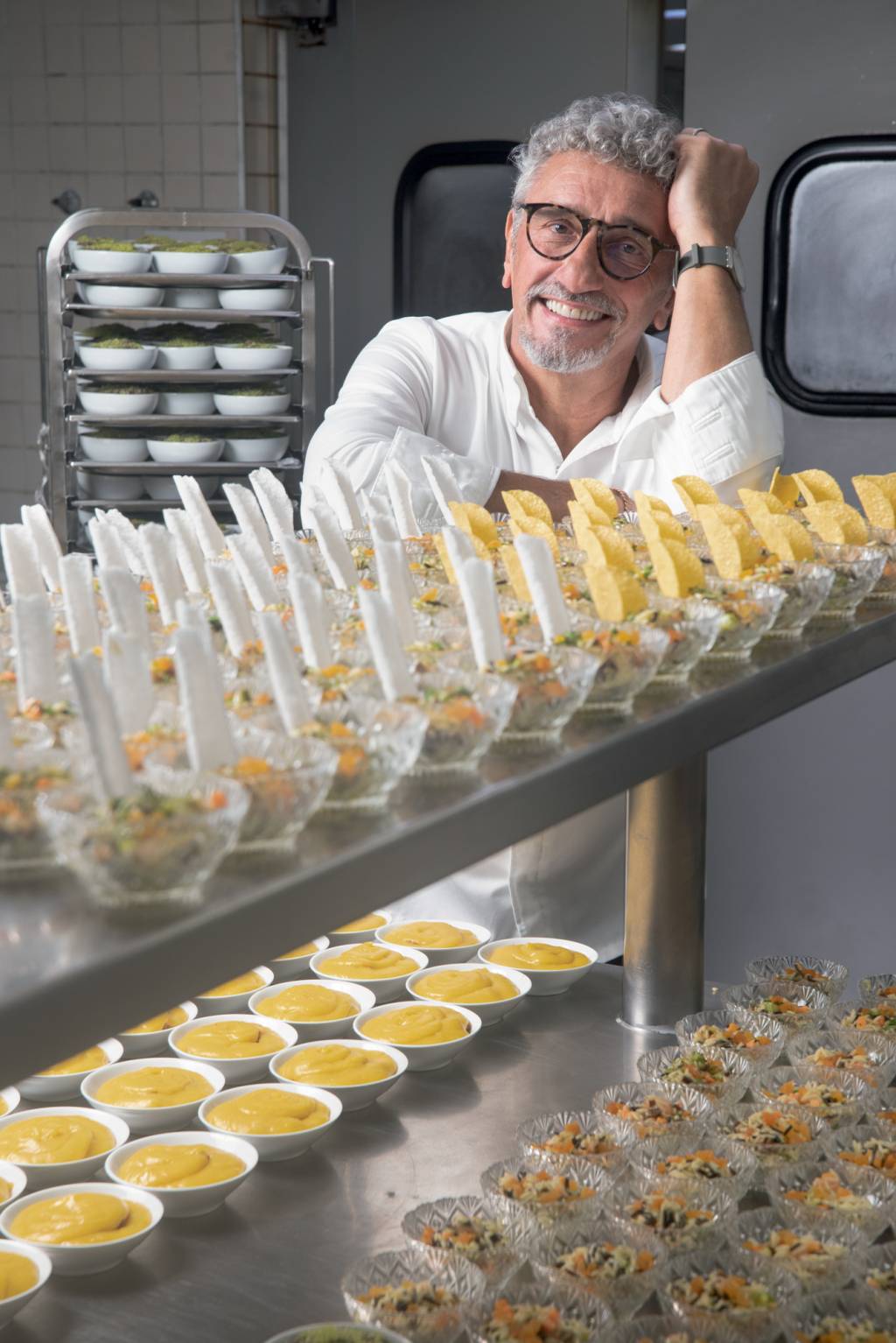 Skye – Emmanuel Bassoleil – Hotel Unique Chef sorrindo para foto com braços apoiados em prateleira metálica linha de produção de prato em cozinha industrial