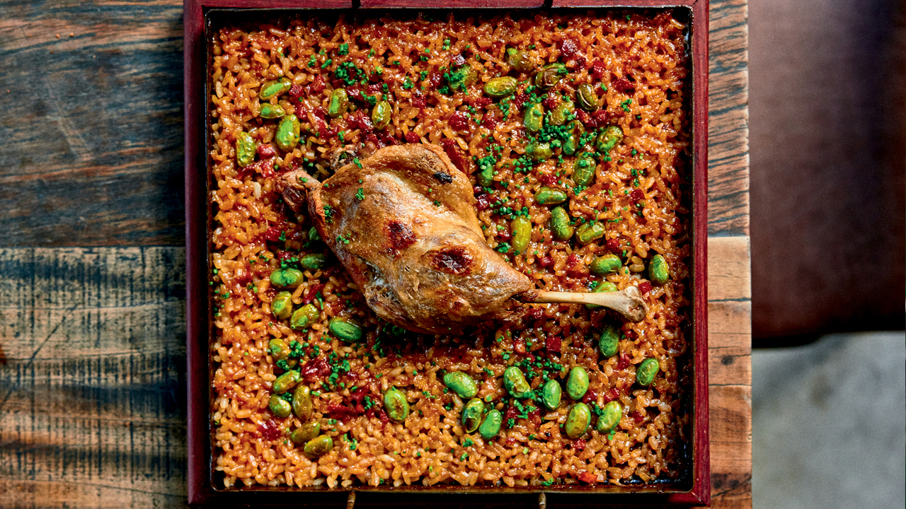 Foto aérea de panela quadrada sobre mesa de madeira com arroz de pato coberto por coxa de pato ao centro.