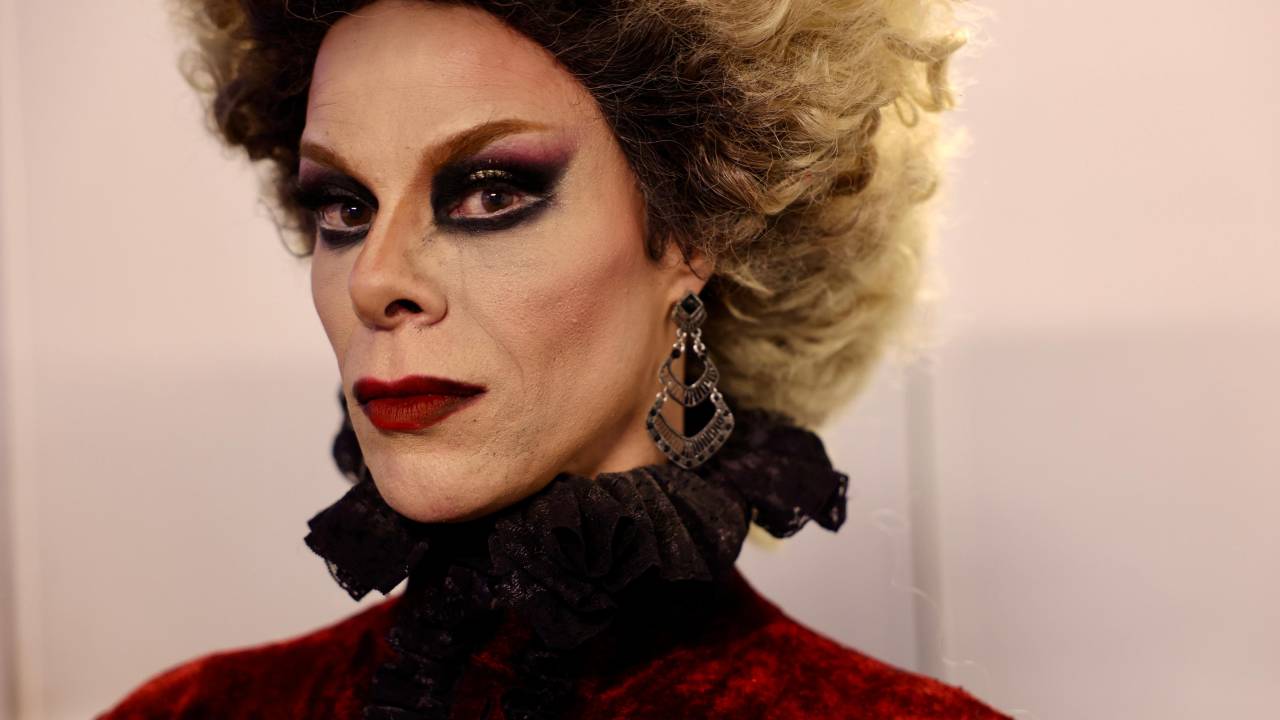 Drag queen com maquiagem preta nos olhos, roupa preta e vermelha até o pescoço e cabelos loiros curtos espichados
