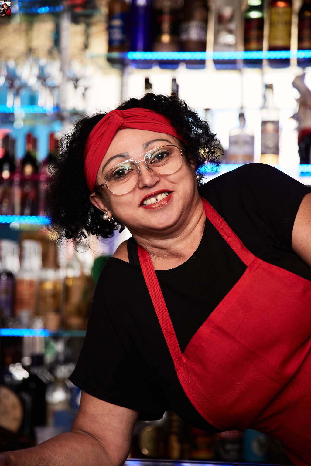 Bar da Bete-Crédito Flávia Faustino-Divulgação (6) Moça branca de óculos grandes de grau sorri apoiada em balcão, veste pano vermelho na cabeça e avental vermelho com camiseta preta