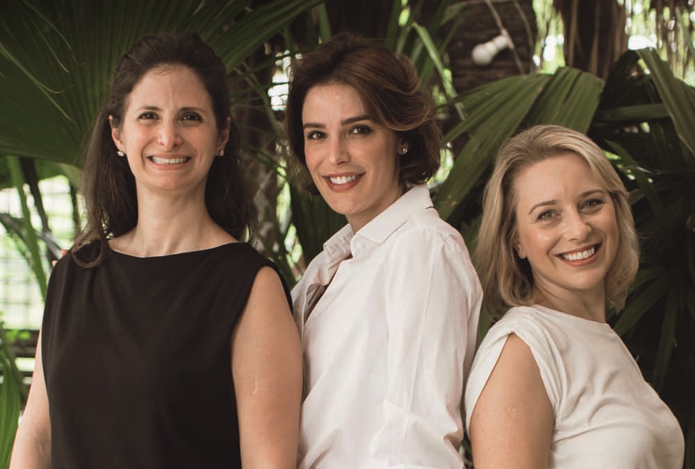 Três mulheres posam lado a lado de pé, sorrindo