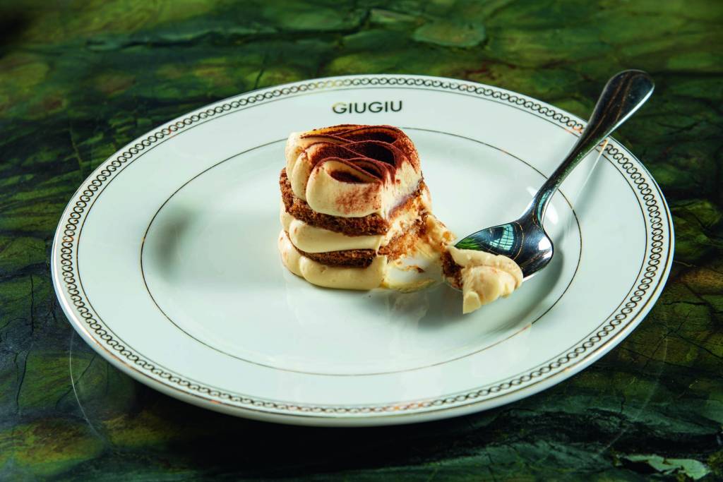 GiuGiu Prato de louça branca com tiramisu cilíndrico com colherada ao lado.