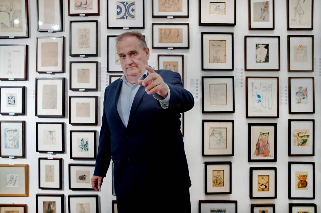 James Lisboa, quadros Aldemir Martins-Crédito Luan Torres-Divulgação (5).jpg Homem corpulento de terno aponta o dedo e posa em frente a série de quadrinhos