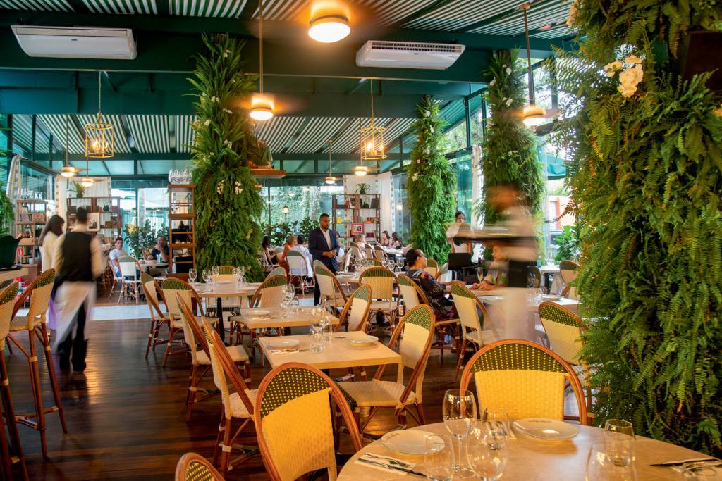 Dia dos Namorados – Les Deux Magots Salão de restaurante com cadeiras de trançado de palha e madeira, piso de taco, luz natural pelas janelas laterais e colunas forradas com samambaias