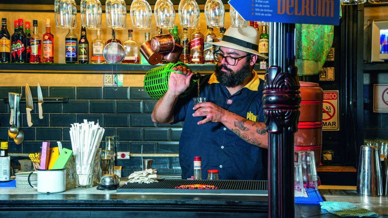 Bartender branco de chapéu panamá e óculos de grau atrás de balcão de bar fazendo drinque.