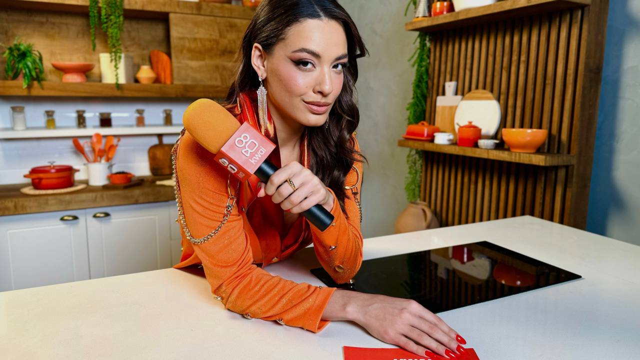 Mulher branca apoiada em balcão branco a sua frente vestida de laranja segurando microfone na mão.