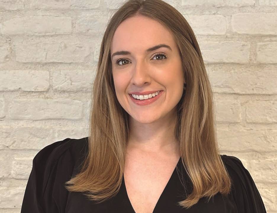 Mulher loira sorri vestindo peça preta e posando em fundo branco de tijolos