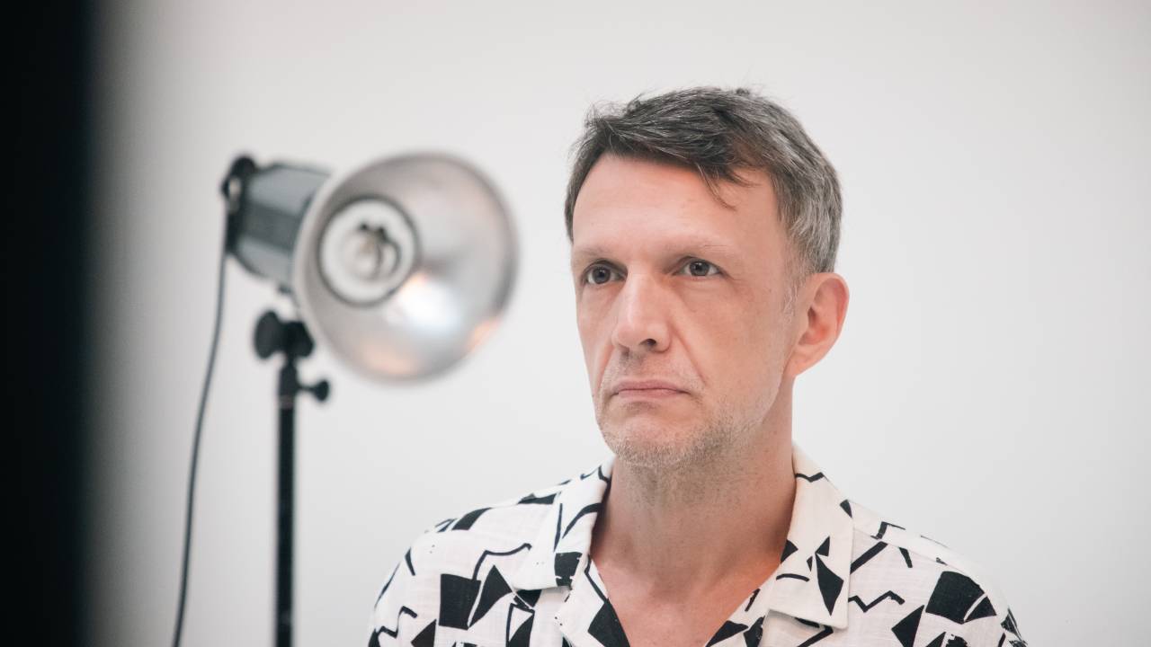 Homem grisalho posa sério vestindo camisa branca e preta