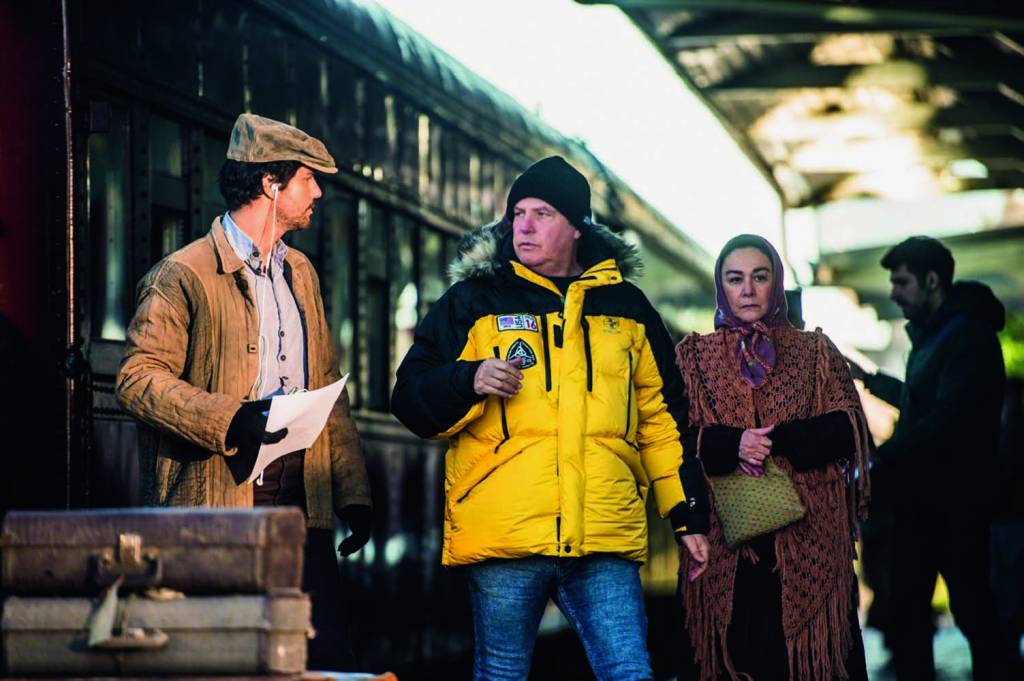 Jayme Monjardim Crédito Arquivo Pessoal (2) Homem branco de casaco amarelo no set de novela com trem de fundo e dois atores