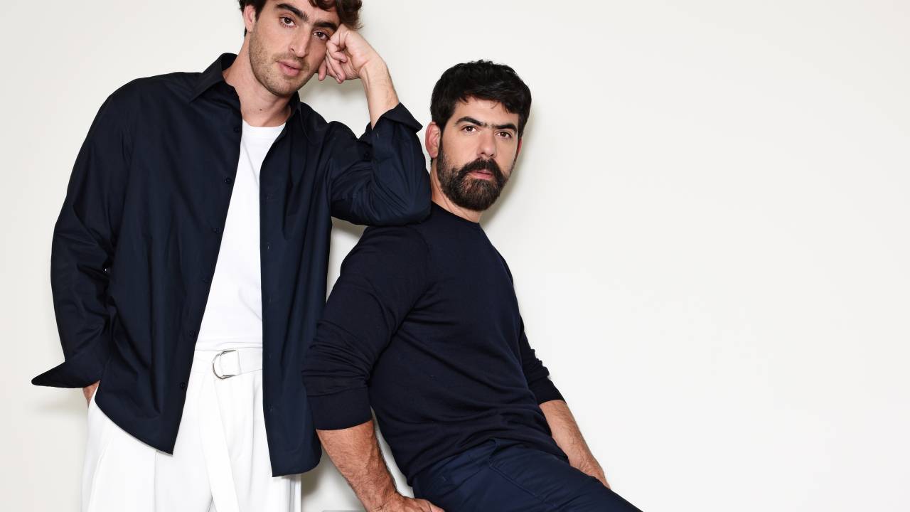 Dois homens brancos posam lado a lado, um de pé, o outro sentado de lado em fundo branco