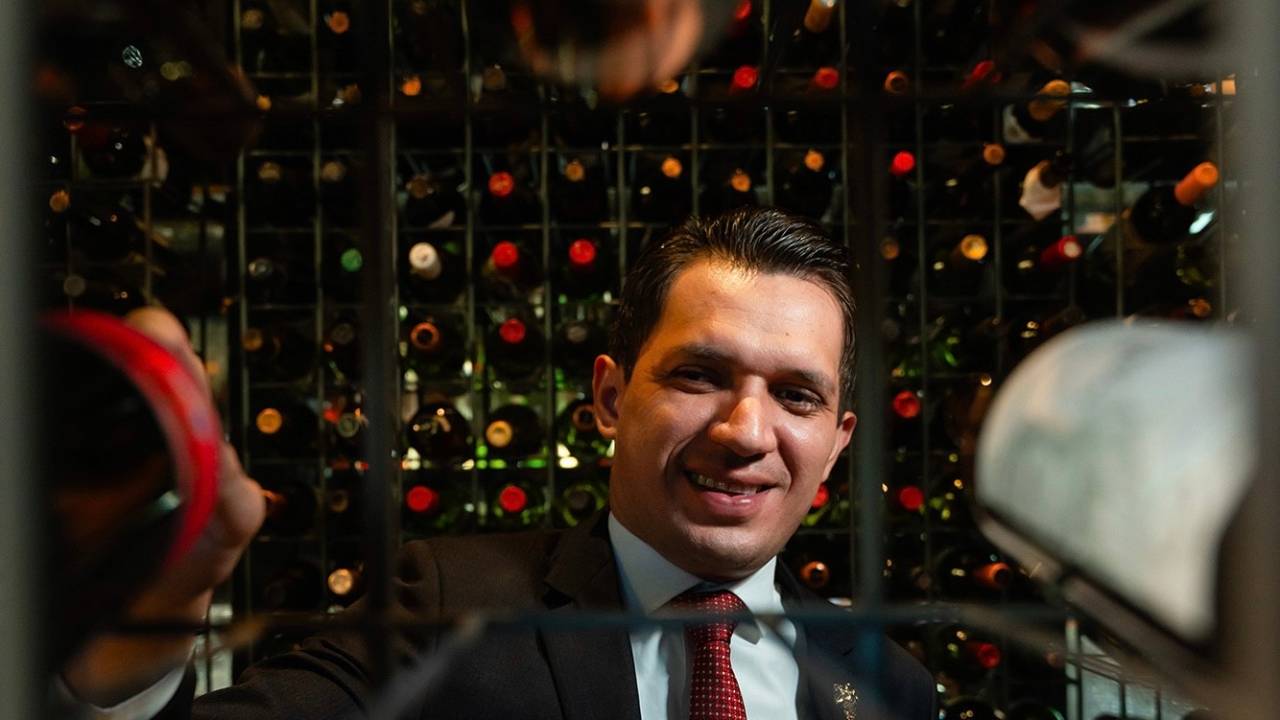 Homem sorrindo em zoom puxando garrafas de vinho de adega.