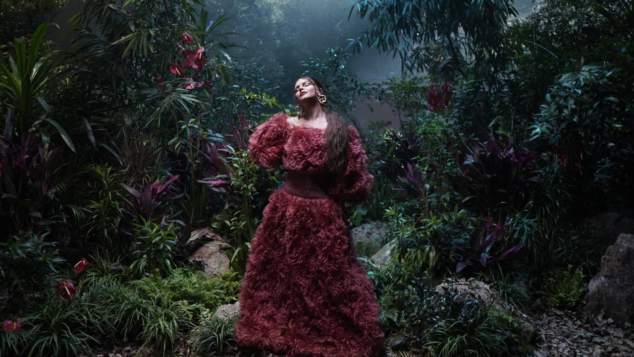 Mulher de vestido vermelho escuro posa em meio à floresta, olhando pra cima