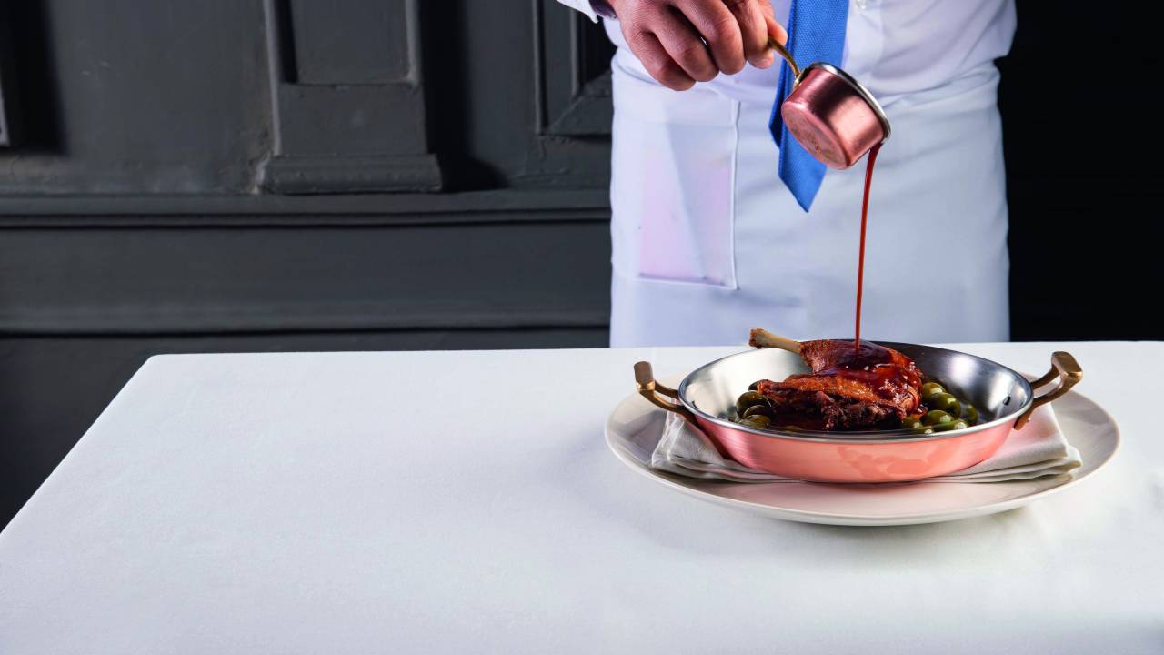 Panelinha de bronze sobre prato de louça branca com coxa de pato dourada sendo coberta por molho numa mesa branca.
