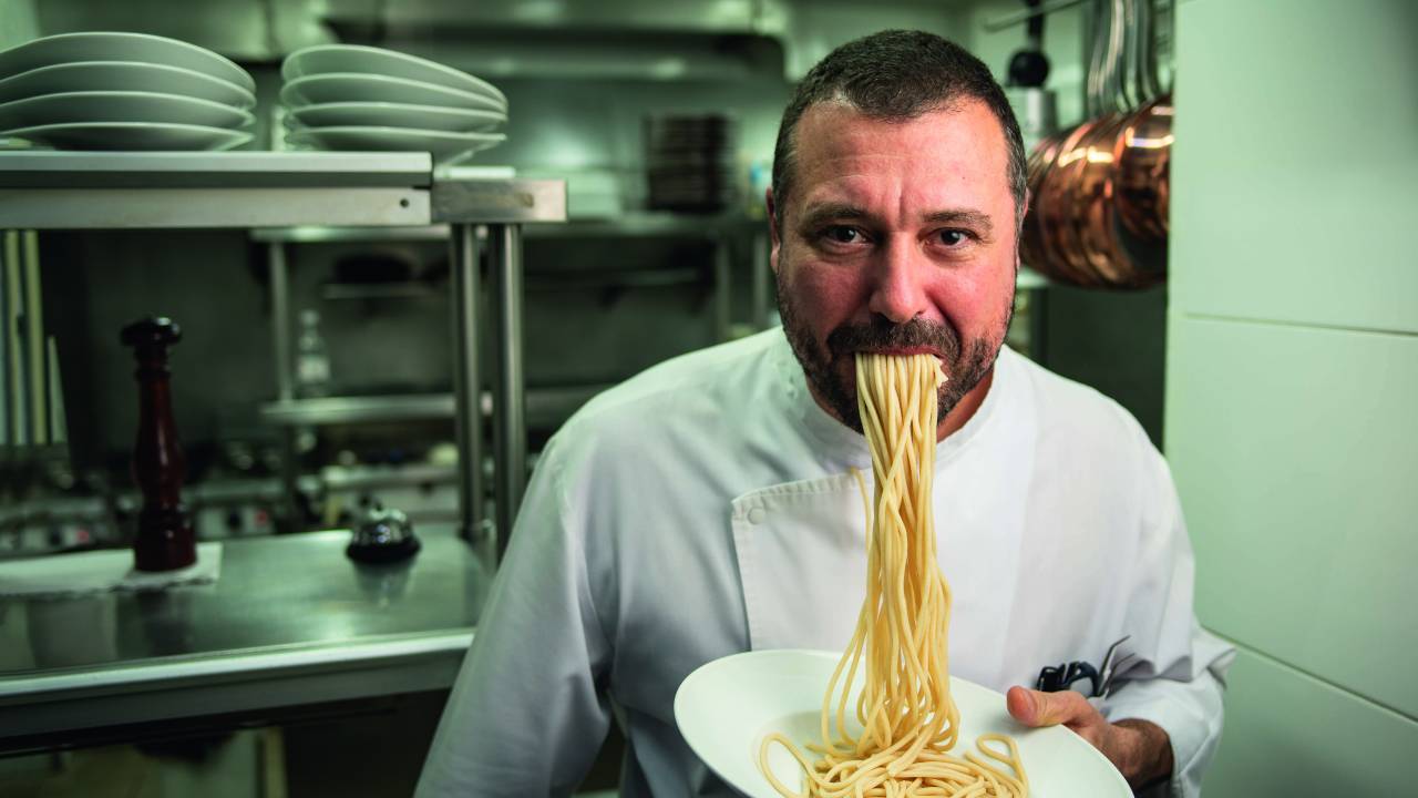 Chef de cozinha de dólmã branco em cozinha industrial com espaguete escorrendo da boca até o o prato de louça branca que está segurando.