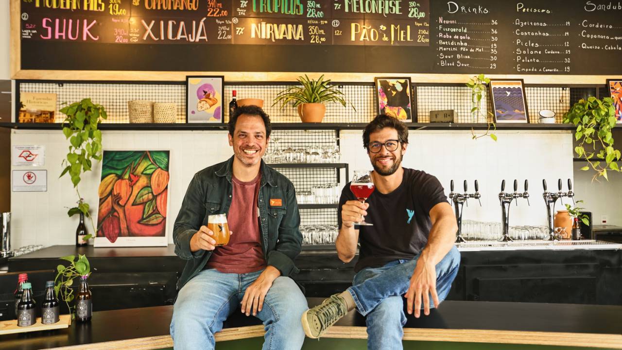 Dois homens sentados em balcão com cerveja na mão e sorrindo