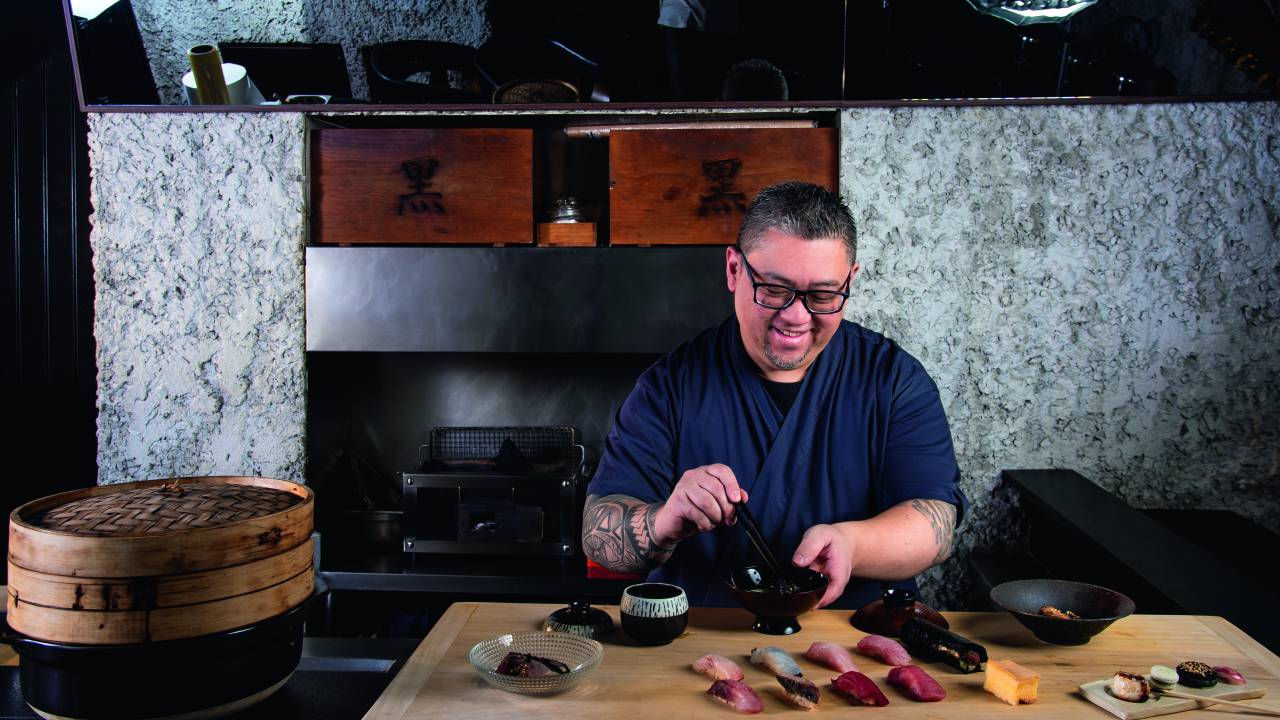 Sushiman segurando hashi preto misturando em tigela preta atrás de bancada de madeira com diferentes sushis e cumbucas por cima.