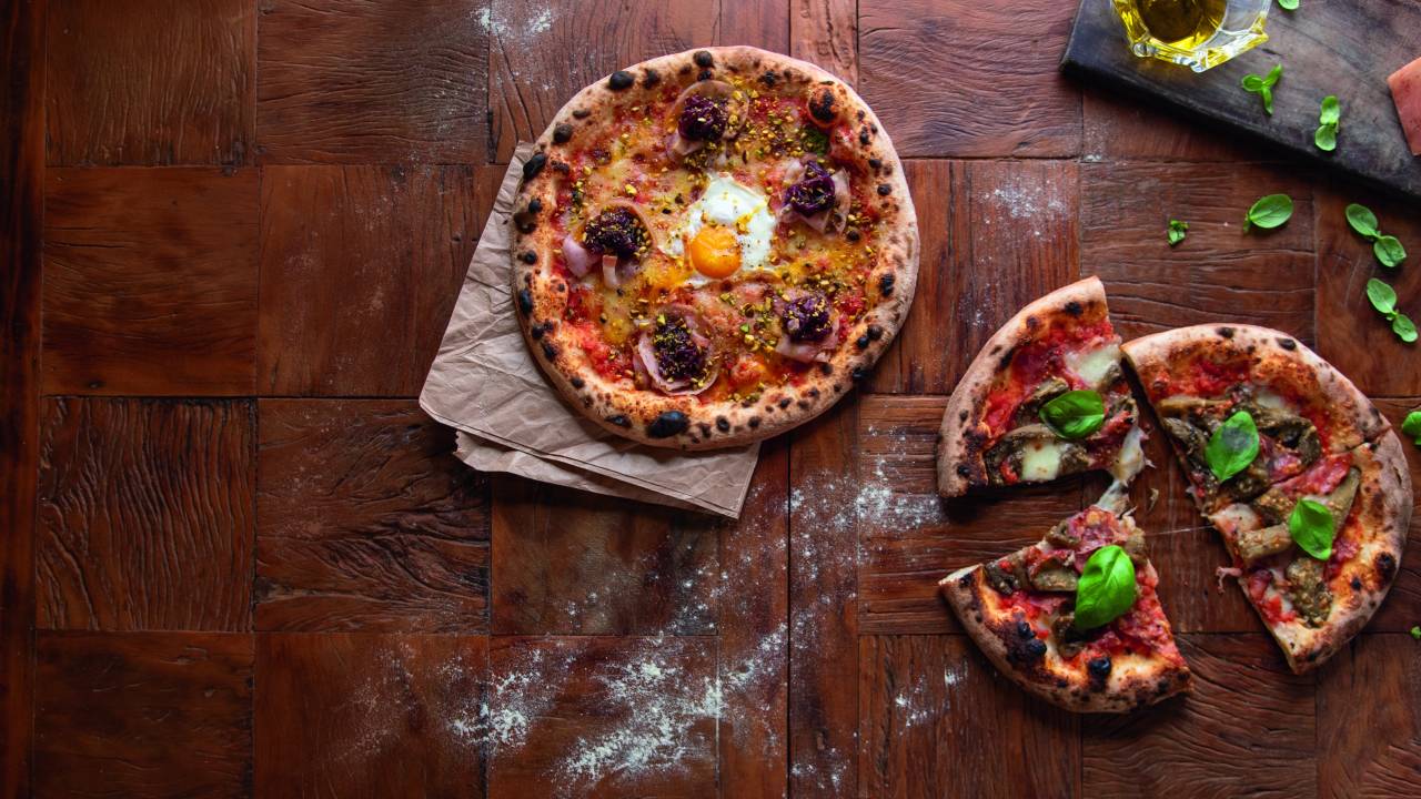 Foto aérea mesa de madeira com duas pizzas por cima. Ao centro, pizza inteira com ovo estalado no meio. No canto inferior à direita, pizza cortada em quatro pedaços coberta por folhas de manjericão.