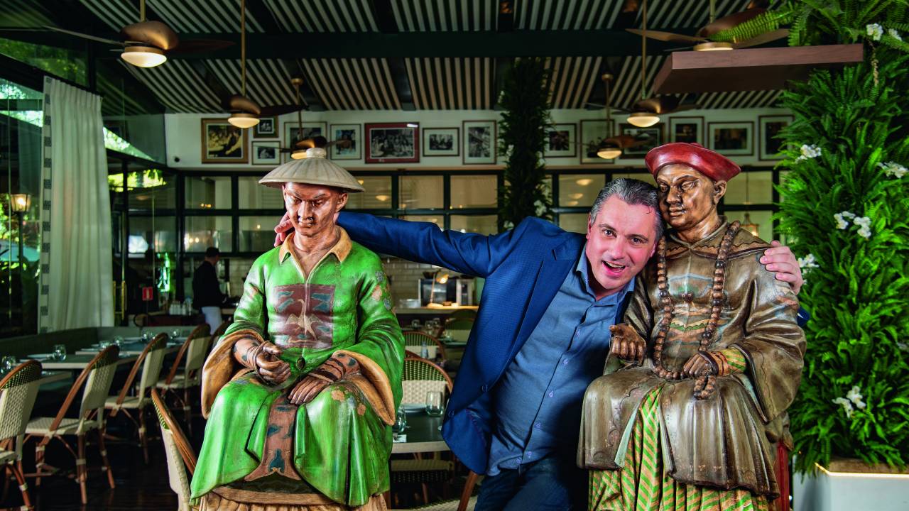 Homem de paletó azul abraçado ao centro com duas estátuas de magos em salão de restaurante.