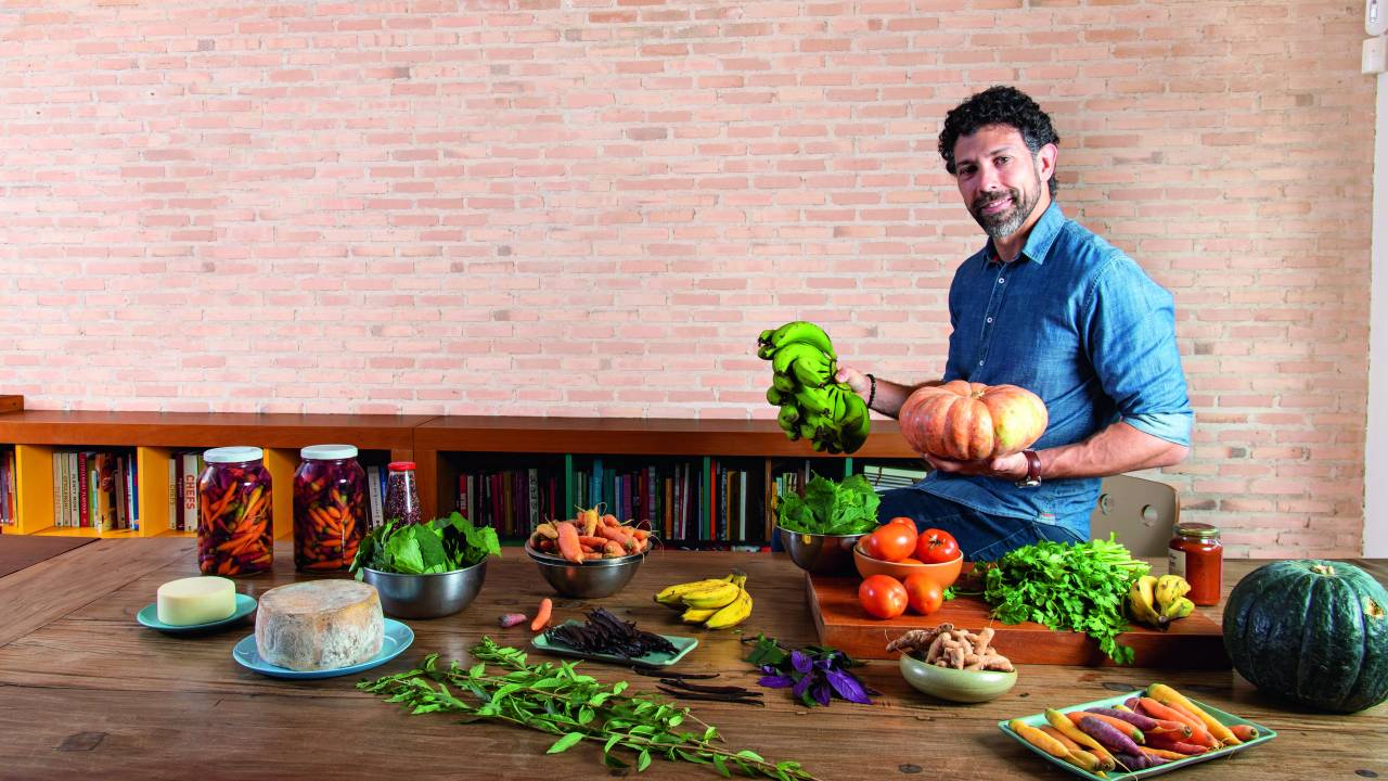 Homem sorrindo para a foto segurando uma abóbora e um cacho de banana atrás de mesa de madeira cheia de verduras e legumes.
