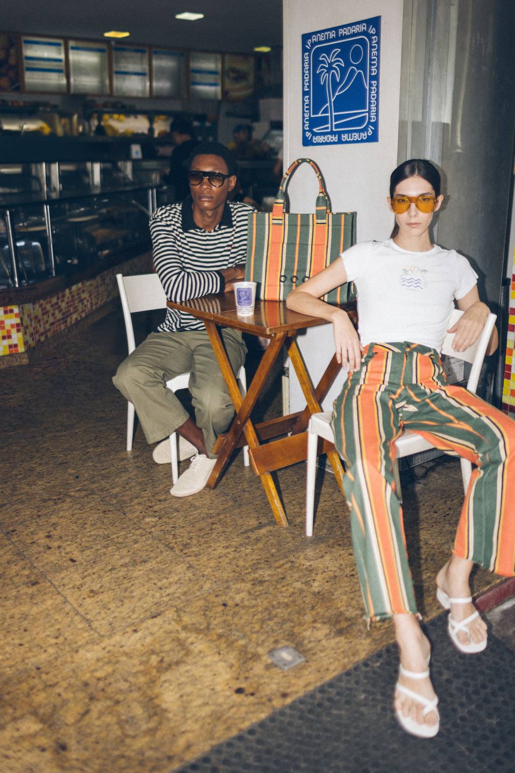 0P8A6716 (1) (1) (2) Moço negro e mulher branca sentados em cadeiras de bar usando peças estampadas, a dela uma calça listrada em tons verde e laranja, ele com blusa de listras pretas e brancas