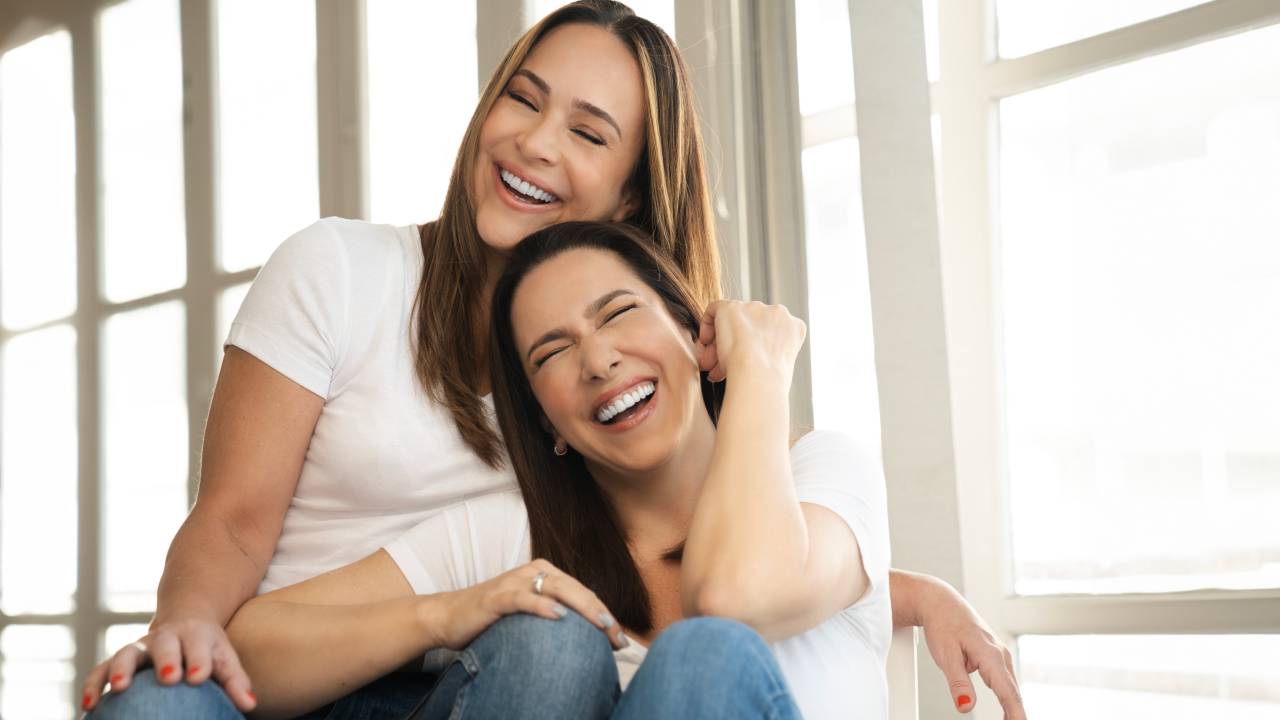 Duas mulheres brancas sorriem abraçadas vestindo jeans e camiseta branca