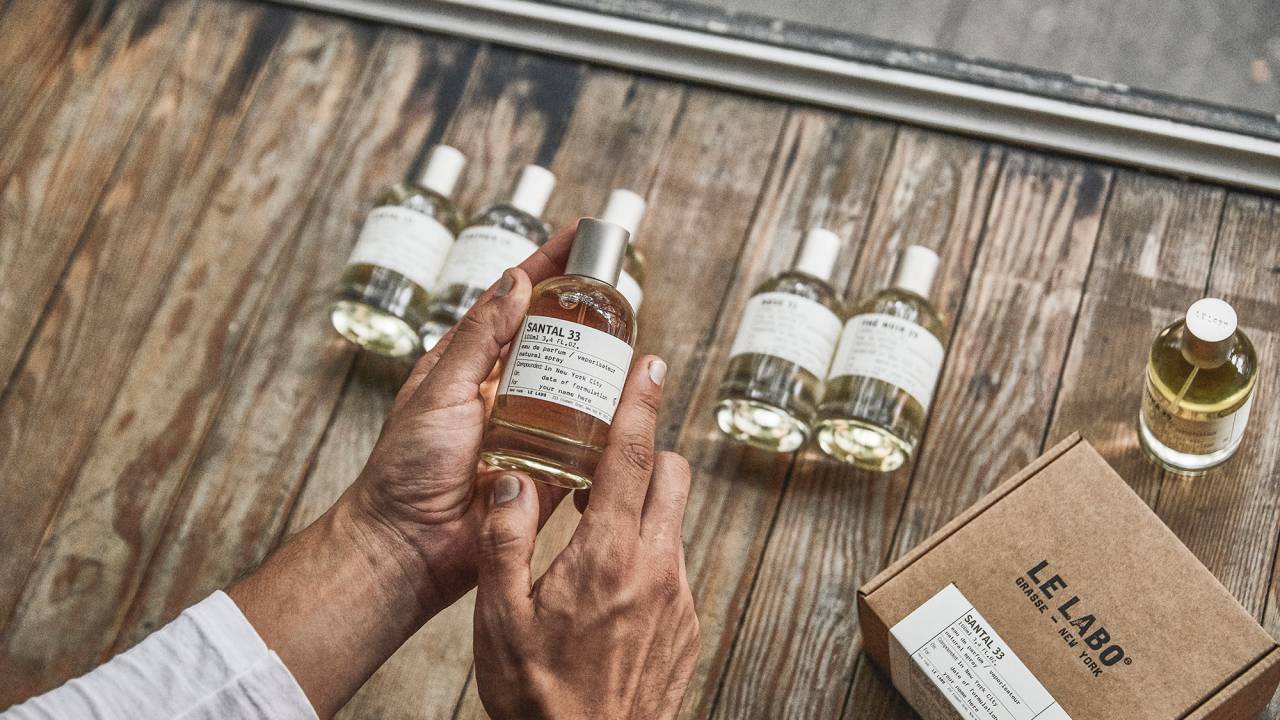 Foto vista de cima com par de mãos manipulando um de 5 frascos de perfume em mesa de madeira