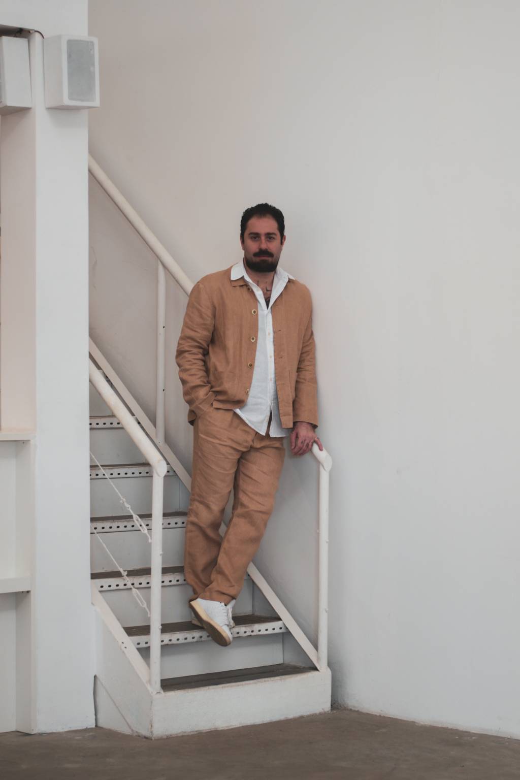 Luis Maluf Crédito Ana Aliaga-Divulgação (3) Homem de barba posa de pé em escada branca, vestindo peça bege e camisa branca por baixo