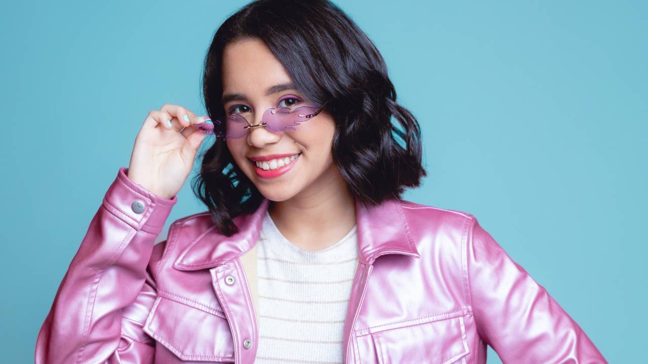 Menina de cabelo curto posa segurando óculos rosa e vestindo jaqueta rosa com calça jeans