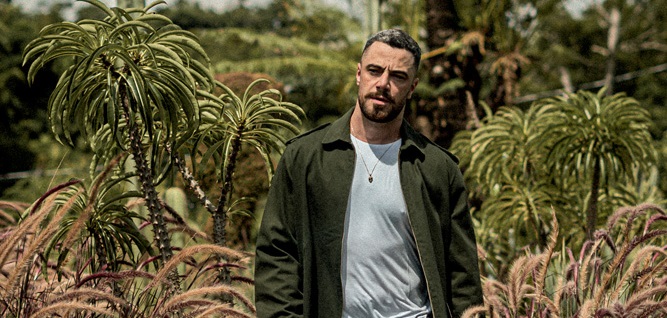 Homem branco de barba anda no meio de uma plantação com jardim ao fundo. Veste jaqueta verde, camiseta clara e jeans clara rasgada. Está segurando um capacete de moto.