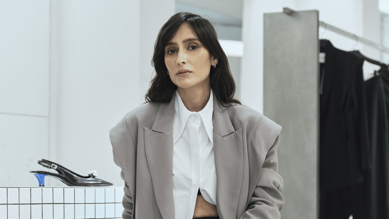 Moça branca de cabelos escuros curtos posa sentada em área de pequenos azulejos brancos com araras e roupas ao fundo. veste blazer cinza e calça preta