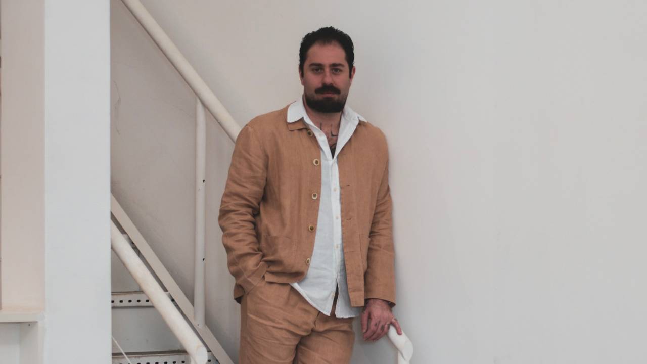 Homem de barba posa de pé em escada branca, vestindo peça bege e camisa branca por baixo
