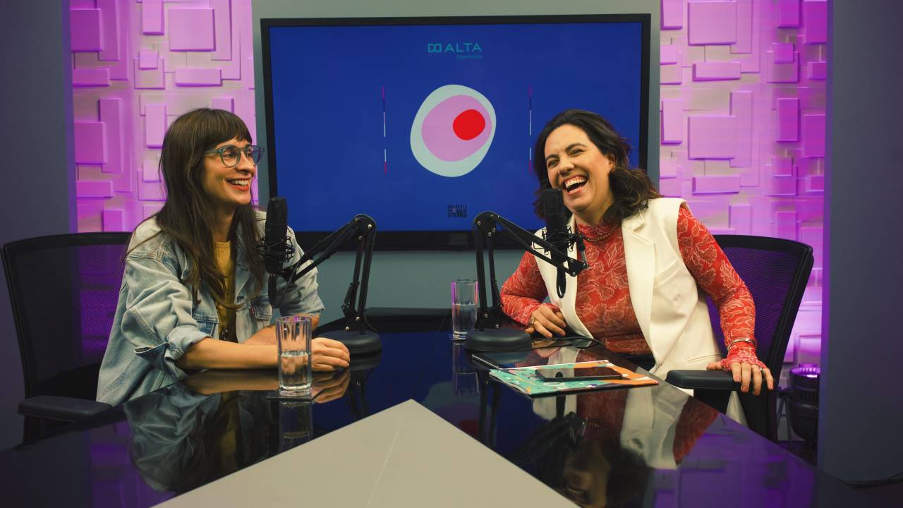 Duas moças em mesa com microfones de podcast e televisão ao fundo