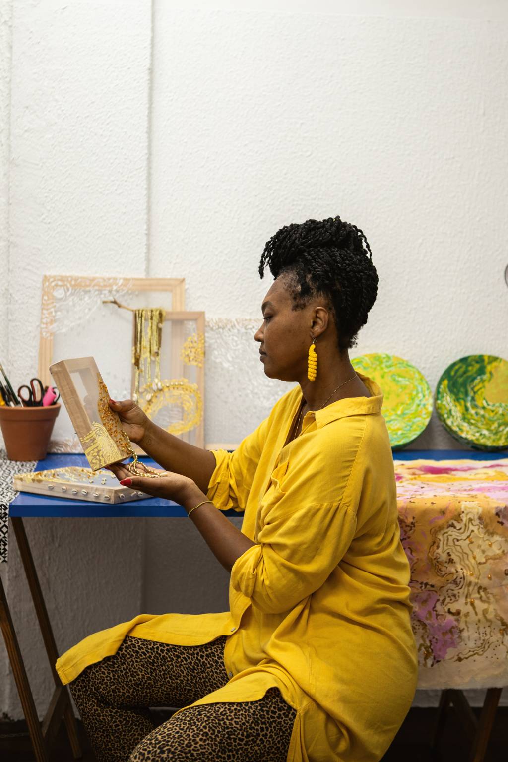 IMG_5287.JPG Mulher negra com roupa amarela sentada de lado pintando em mesa