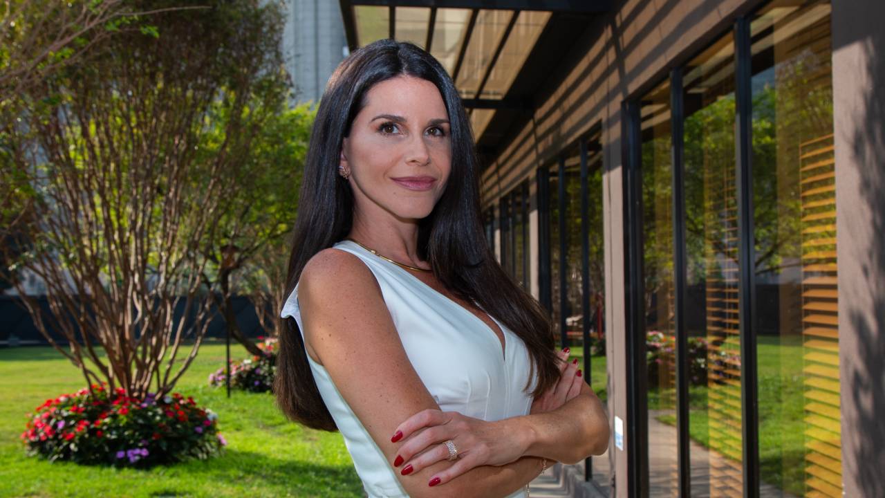 Mulher de braços cruzados veste peça branca em área aberta com jardim