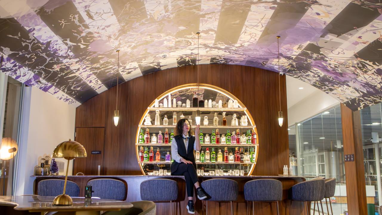 Moça de terninho e gravata borboleta senta em balcão de bar com prateleiras iluminadas ao fundo