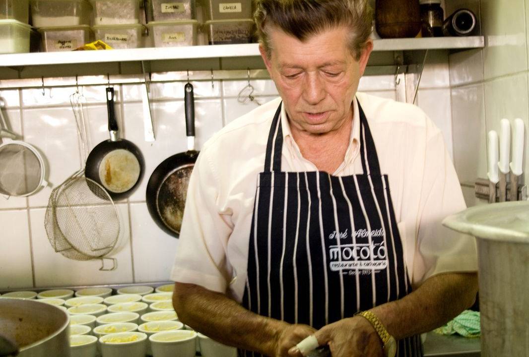 Um homem de avental de listras brancas e pretas em um cozinha com uma panela de pressão nas mãos