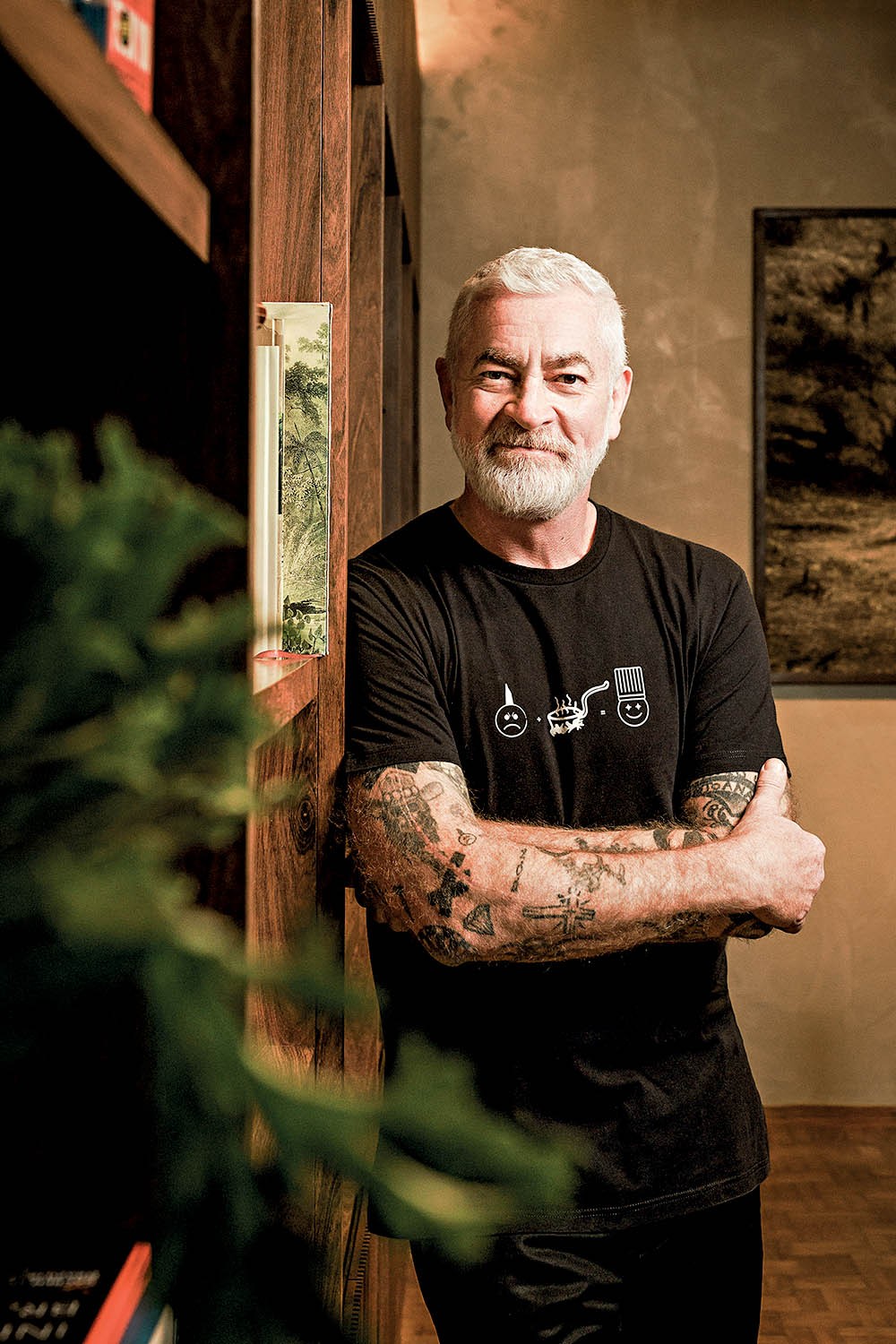 Homem branco de barba e cabelo grisalhos, sorrindo, com bra&ccedil;os tatuados cruzados, vestindo camiseta preta com estampas de emojis e um chap&eacute;u de chef, encostado em uma estante de madeira com livros e plantas
