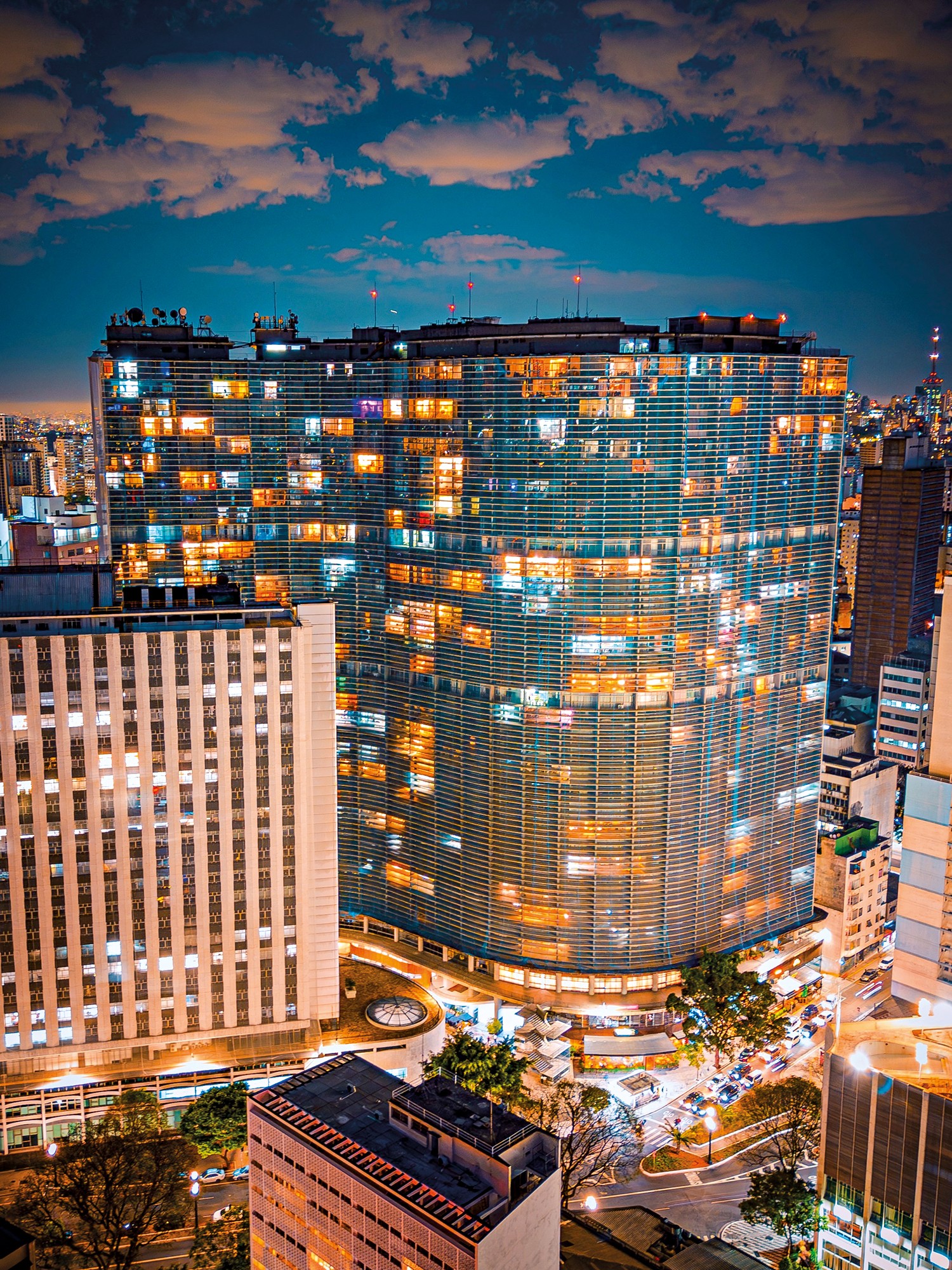 Vista aérea noturna de arranha-céus em São Paulo, com um edifício curvo de fachada azul e janelas iluminadas em tons de laranja e branco. À esquerda, um prédio branco retangular também iluminado. Abaixo, ruas com carros e árvores, sob um céu azul escuro com nuvens claras