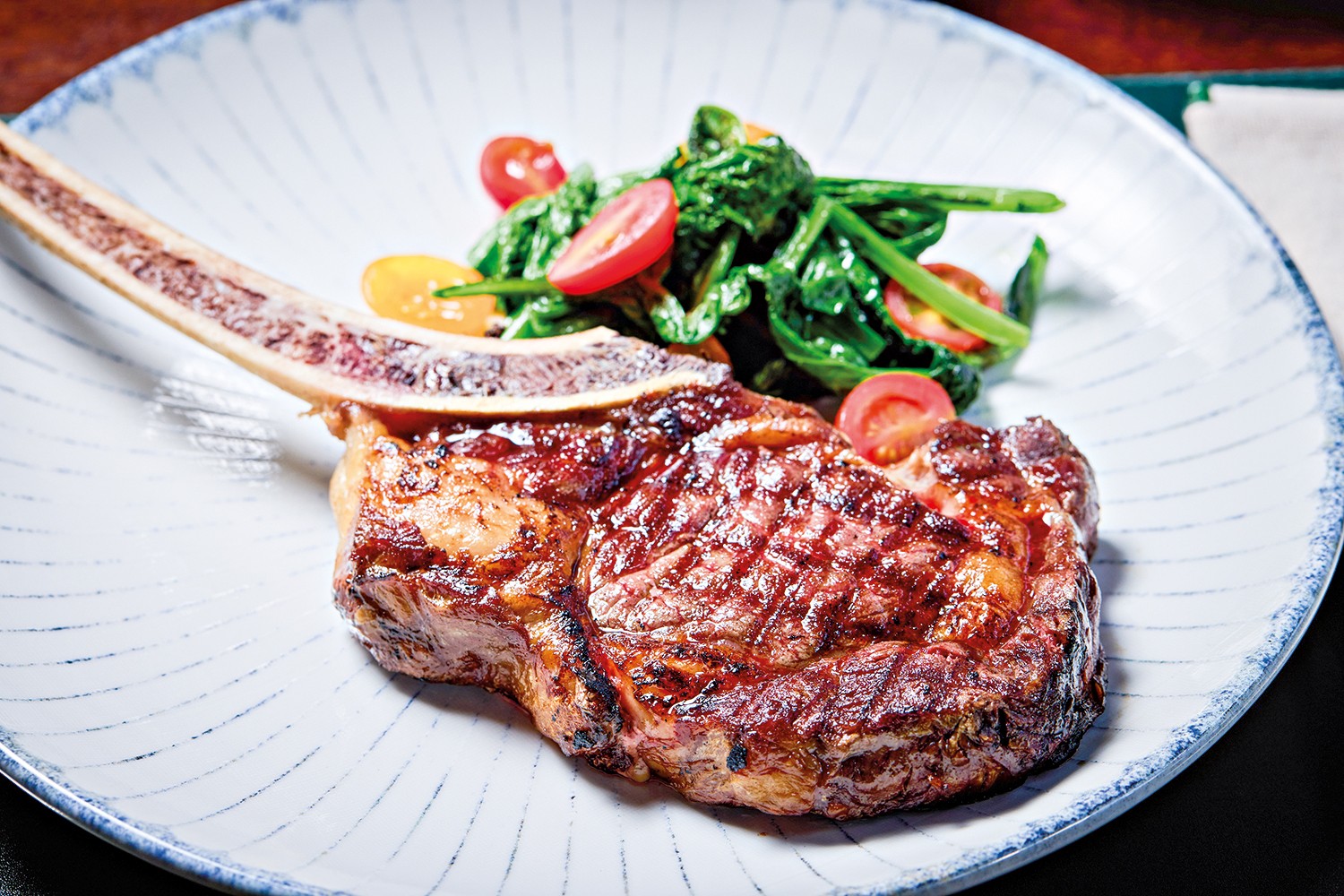 Bife tomahawk grelhado com osso, suculento e com marcas de grelha, servido em prato branco com borda azul e linhas finas. Ao lado, um acompanhamento de folhas verdes escuras, tomates cereja cortados ao meio e piment&otilde;es amarelos