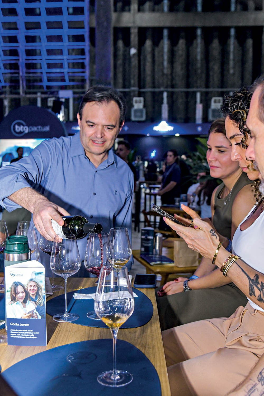 Homem de camisa azul serve vinho tinto em ta&ccedil;as, enquanto tr&ecirc;s mulheres observam. Uma delas, com tatuagens e pulseiras douradas, segura um celular. H&aacute; um folheto Conta Jovem do BTG Pactual na mesa. O ambiente &eacute; um evento noturno com ilumina&ccedil;&atilde;o azulada