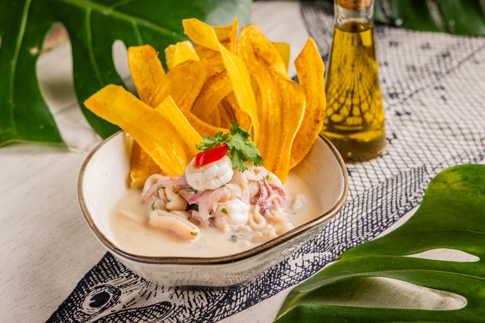 Ceviche de camar&atilde;o e lula em molho cremoso, guarnecido com coentro e pimenta vermelha, acompanhado de chips de banana-da-terra crocantes. Uma garrafa de azeite e folhas verdes tropicais decoram a mesa com toalha estampada