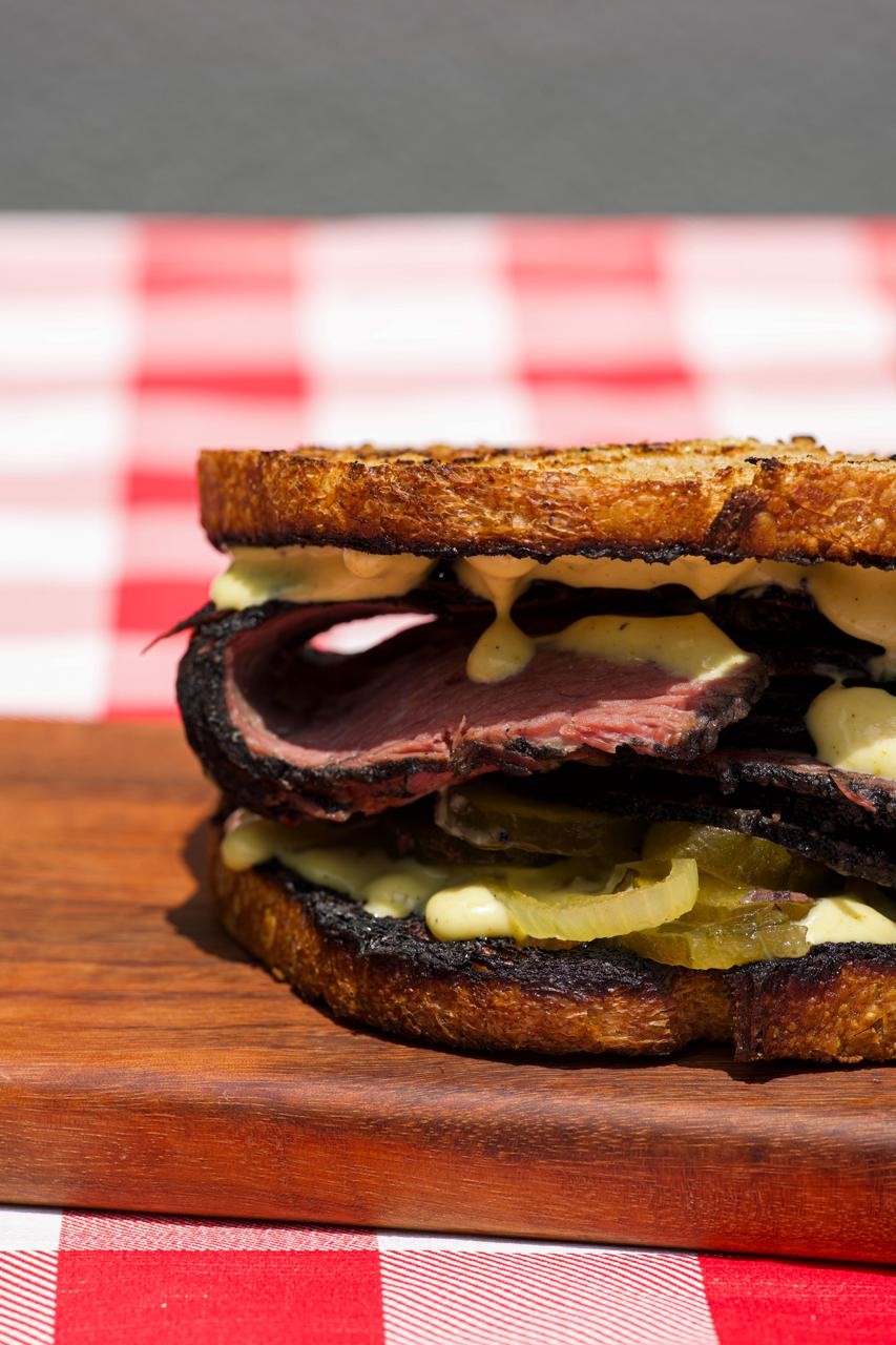 Sandu&iacute;che de pastrami com fatias de carne vermelha, picles e molho cremoso, servido em p&atilde;o torrado sobre uma t&aacute;bua de madeira, com toalha xadrez vermelha e branca ao fundo