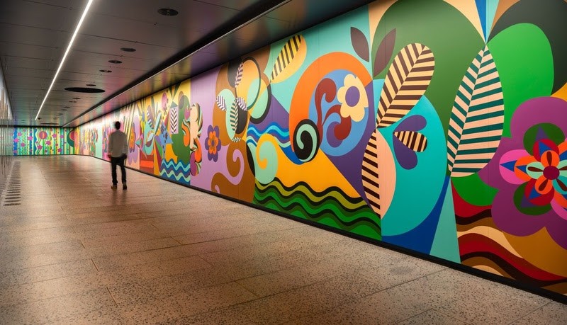 Homem de costas em um corredor iluminado, observando um mural vibrante com formas abstratas e cores como azul, laranja, verde e roxo, que se estende por toda a parede lateral
